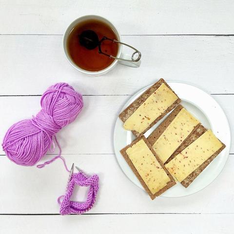 Flat lay view of a plate with 4 crackers with cheese, a large cup of tea, and a small knitting project on a tiny circular needle with its lilac ball of yarn on a white plank background.