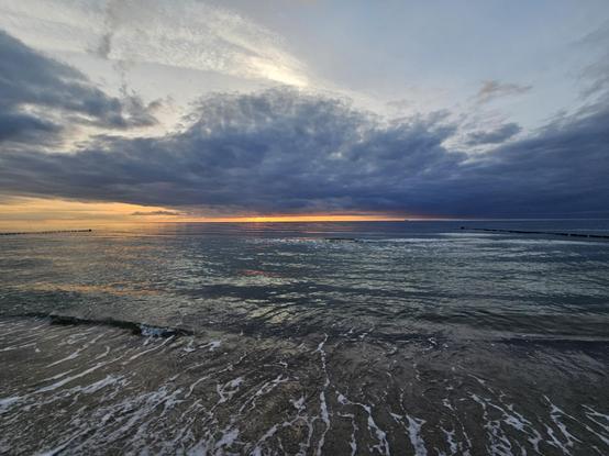 Auf diesem Foto habe ich den Sonnenuntergang am Meer festgehalten. Am Horizont leuchtet ein schmaler goldfarbener Steifen, darüber eine große, dunkle Wolke, die nach vorn hin in ein helleres Blau übergeht. Die Meeresoberfläche ist leicht gewellt und spiegelt die Farben des Himmels wieder.