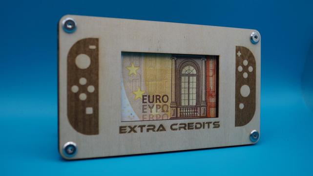 Holzrahmen in Form einer Spielkonsole mit Gravur „Extra Credits“, mittig ein sichtbarer Geldschein im „Bildschirm“.