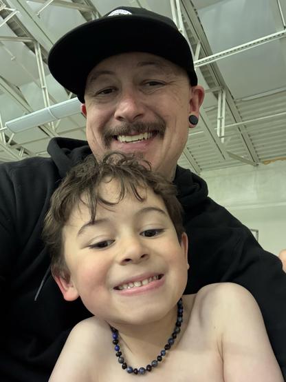 A man with a mustache and earrings smiles widely at the camera, holding a young boy who is grinning and wearing a beaded necklace. The boy is shirtless, and they are both indoors, with a ceiling visible above them.