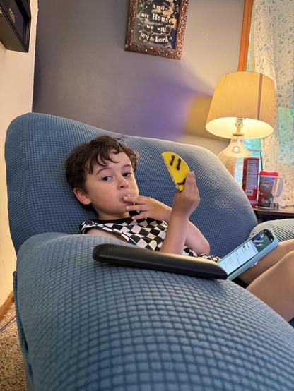 A young child sits on a blue couch, holding a yellow object resembling a banana while looking at the camera. The child is dressed in a black and white checkered outfit and appears to be engaged with a mobile device resting on the couch. 
