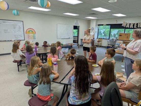 A group of children sits at a table in a classroom, attentively listening to an instructor. The room features a whiteboard, colorful decorations, and large windows with greenery visible outside. Some children are enjoying snacks.