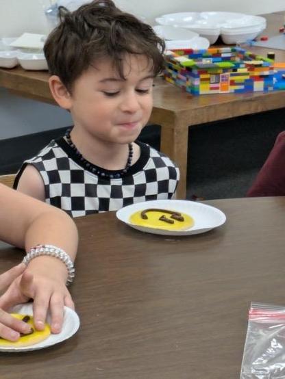 A young child with curly hair is sitting at a table, looking at a plate with a yellow icing decoration shaped like the number "3" and dark candies. There are other plates and colorful building blocks in the background.