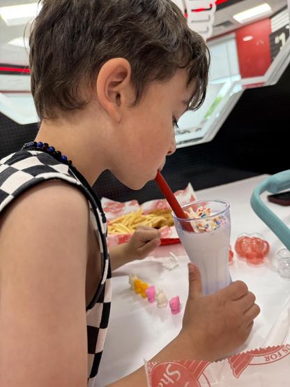 A child with short hair is sipping a milkshake through a red straw. The shake is topped with colorful sprinkles. In front of the child, there is a tray with fries and small, colorful gummy candies. The background features a modern diner setting