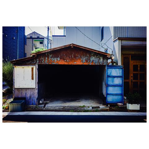 This image captures a dilapidated one-car garage sandwiched tightly between more modern residential buildings in Nagoya. The roof is rusted to an almost painterly orange, while the right-hand wall is an oxidized blue metal panel that looks like it’s borrowed from a shipyard. The gaping blackness inside offers no visual cues—just pure, uninterrupted darkness. Two mismatched planters try their best to lend charm, but the overwhelming impression is one of beautiful abandonment in broad daylight.