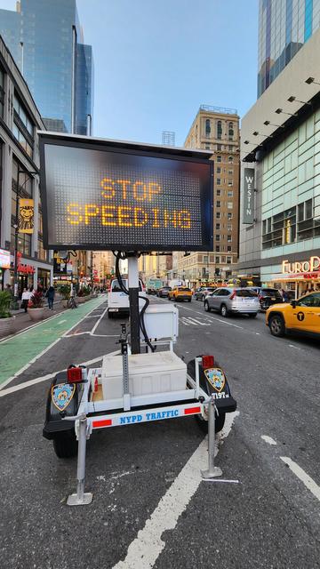 A movable NYPD sign board programmed to read STOP SPEEDING, positioned between a green painted bike lane and auto traffic on a busy Manhattan avenue.
