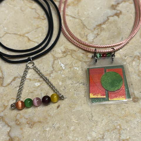 On cream marble background two cords, one black the other pink, coiled above their pendants. Black has five beads (orange, green, pink, purple, yellow on a wire) hanging in a line forming the base of a triangle between two chains. The pink pendant is laminated coloured paper (three layers:green square, two vertical orange rectangles, overlayed with a smaller green circle) at top three small beads (green, orange, green)