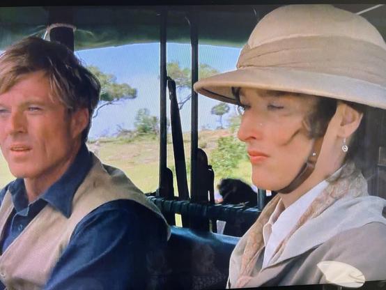 A man and a woman are sitting in a vintage vehicle, likely on a safari. The man, dressed in a dark shirt and vest, appears focused on the landscape outside. The woman, wearing a light-colored hat and jacket, looks contemplative.
