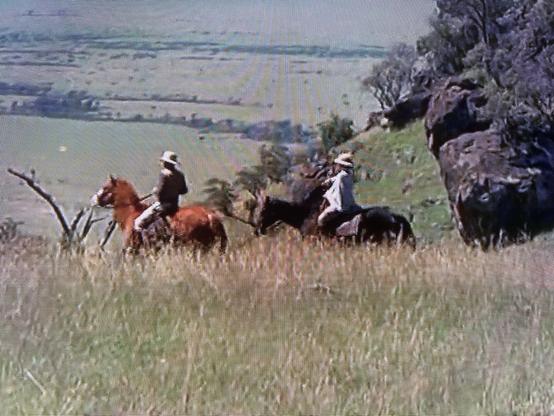 Two horseback riders are seen on a grassy hillside, with greenery and rocky terrain in the background. The riders are wearing hats and appear to be enjoying the open landscape.