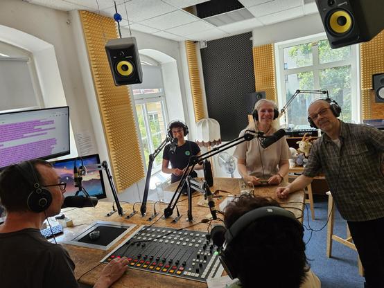 Fünf Menschen sitzen in einem Aufnahmestudio. Sie haben Kopfhörer auf. Vor jeder Person steht ein Mikrophon. In der Mitte befindet sich ein Tisch mit Bildschirmen, Knöpfen und Lautstärkereglern.