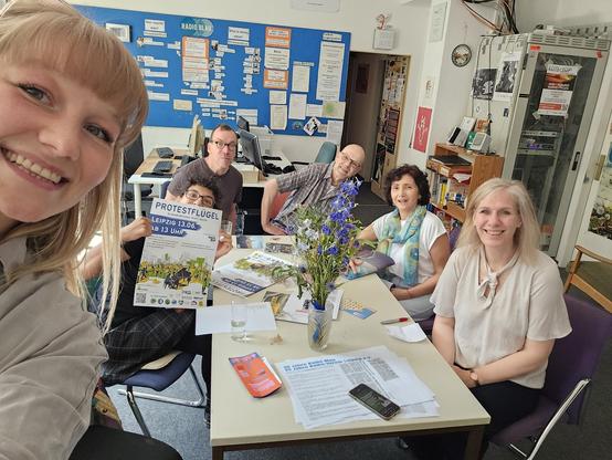 Sechs Menschen sitzen gemeinsam an einem Tisch. Sie lächeln alle in die Kamera. Eine Frau mit Brille hält ein ProtestFlügel-Plakat hoch. Auf dem Tisch liegen weitere Plakate und Flyer um eine Blumenvase herum.