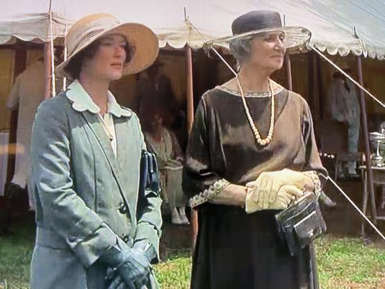 Two women are standing side by side in front of a tent. One woman wears a light green coat and a wide-brimmed straw hat, while the other wears a dark dress and a wide-brimmed black hat. 