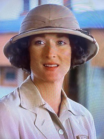 A woman with curly hair is wearing a light-colored safari hat and a matching shirt. She appears to be smiling, with a warm expression, in a setting that suggests an outdoor or adventure theme.