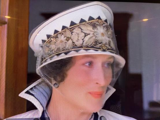 A woman in a stylish, vintage hat with a decorative band is depicted. She has light skin and is wearing a chic outfit with a high collar. The background features wooden architectural elements, suggesting an indoor setting.
