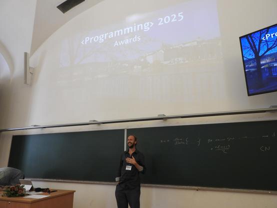 A photo of general chair Tomas Petricek at the <Programming> 2025 award ceremony. Tomas is standing in the lower part of the picture in front of a chalkboard, smiling at the audience. Above him is a projected slide with a photo of a river in Prague at night with text saying "<Programming> 2025 Awards".
