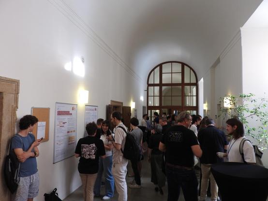 A photo of the first round of the student research competition at <Programming> 2025. There is a hallway with 7 SRC posters pinned to the walls. Groups of people are standing around the posters; the students explaining their research to the audience.