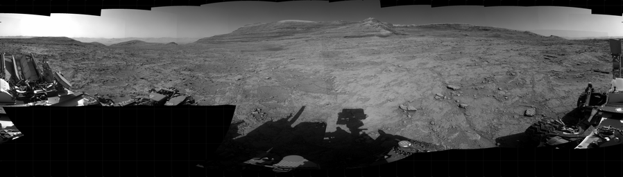A black and white 31 images mosaic capturing a Martian landscape, with the shadow of the Curiosity rover projected at center in the foreground