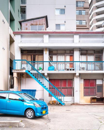 Das Bild zeigt die Rückseite eines älteren zweistöckigen Wohngebäudes mit einem sichtbaren Außenaufgang. Die Architektur wirkt einfach und funktional. Die untere Etage besteht aus mehreren Türen und Fenstern mit rotbraunen Rollläden oder Jalousien. Die Fassade ist größtenteils grau, teils verfärbt oder leicht fleckig, was auf das Alter und eine gewisse Vernachlässigung hinweist.

Eine außenliegende Metalltreppe, die in einem leuchtenden Blau gestrichen ist, führt schräg von unten links nach oben rechts in das obere Stockwerk. Oben verläuft ein durchgängiger schmaler Balkon mit einem dazu passenden blauen Geländer.

Auf dem Balkon sind mehrere Alltagsgegenstände abgestellt, darunter blaue Plastiksäcke, eventuell mit Müll oder Recyclingmaterialien, und einige Haushaltsartikel. Vor den Fenstern des oberen Stocks hängen unterschiedliche Vorhänge, teils durchsichtig, teils blickdicht in Weiß und Orange.

Links unten im Bild steht ein kleines, leuchtend blaues Auto auf einem offenen Parkplatz. Es hat ein gelbes Kennzeichen, was auf eine japanische Herkunft schließen lässt (gelbe Nummernschilder werden in Japan für Kleinfahrzeuge, sogenannte „Kei-Cars“, verwendet).

Im Hintergrund ragen größere und modernere Hochhäuser empor, die deutlich höher und gepflegter wirken als das Gebäude im Vordergrund. Auf dem Dach des älteren Gebäudes befindet sich noch eine kleine zusätzliche Holzkonstruktion mit Wellblechdach – möglicherweise ein Abstellraum oder ein improvisiertes Dachgeschoss.