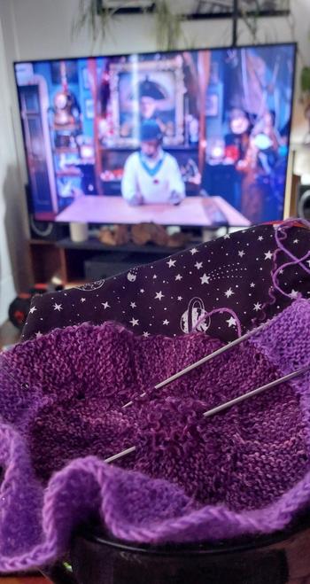A circle of purple knitting in a black knitting bowl in the foreground, and an out of focus tv in the background, showing a man in cricket whites in a room full of things, an episode from series 18 of Taskmaster.
