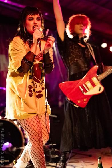 A female singer with edgy fashion, wearing a yellow oversized t-shirt and fishnet stockings, performs with a microphone. In the background, another performer with bright red hair raises their arm, holding a red guitar. The setting is a lively music venue.