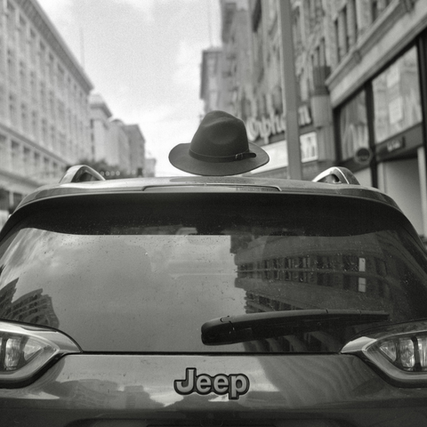 A black and white film photo. A Jeep SUV is wearing a hat in an attempt to cover a bald spot in downtown Los Angeles, California. Sunday, May 18, 2025.