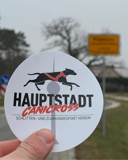 Eine Hand hält den Aufkleber des Vereins "Hauptstadt Canicross" vor die Kamera. Im Hintergrund ist unscharf ein Ortsschild zu sehen.