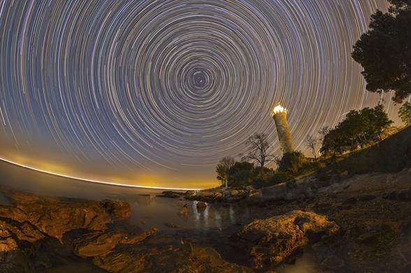 Long-exposure night photograph of Savudrija lighthouse on the Adriatic coast, with circular star trails filling the sky. The lighthouse stands illuminated near the bottom left, while the stars form concentric arcs centered around Polaris near the top center of the image. The trails create a sense of rotation, emphasizing Earth's movement. The image has a serene and mesmerizing quality, blending coastal architecture with cosmic motion.