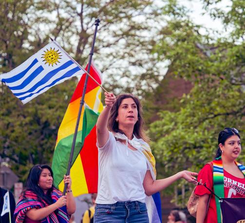 A woman with a white blouse waves a flag with blue and white stripes and a sun in the field while another woman behind her holds a much bigger red/yellow/green striped flag