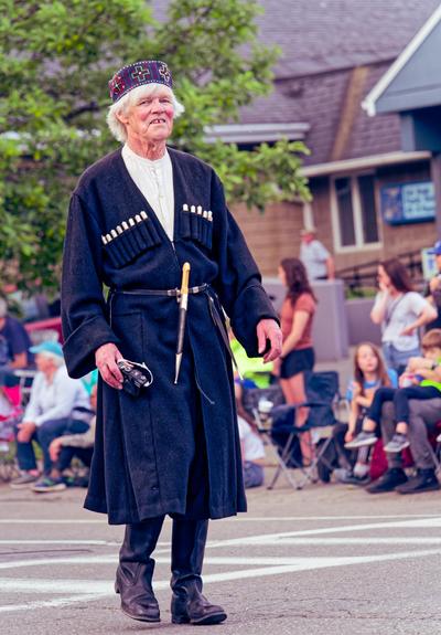 A man in a long blue robe, white hair and cylindrical cap with plus sign markings walks pass sidewalk with crowd sitting on street behind him.
