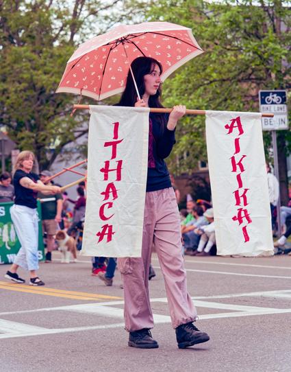 Woman in dark blue shirt and light pinkish brown poants holds a pink umbrella with white markings and a light colored border holds a rod with banners that sai ITHACA AIKIKAI 