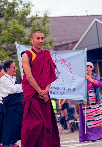 Man in yellow and magenta robe walks in from of sign with tibetan characters held by two people behind him