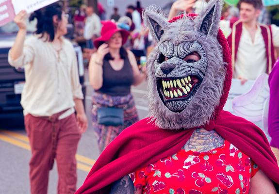 Woman in march with wolf mask with upright ears and large teeth wears a red hood hood and also a red blouse with groups of cherries and slices of cherry pie with several marchers behind, also the grille of a truck and the crowd behind.