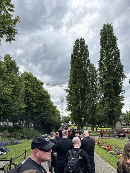 A line of people dressed in black is seen in a park, with tall trees and overcast skies in the background. There are colorful flowers and some park benches visible, along with a construction crane in the distance.