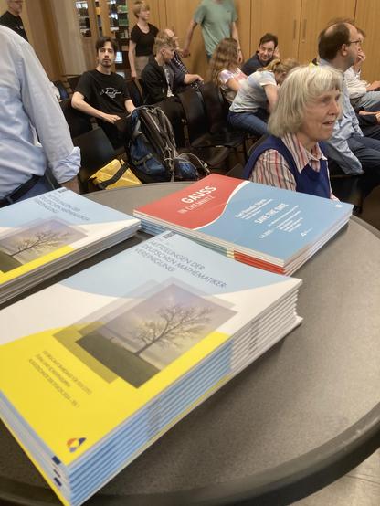 A stack of colorful brochures titled "Gauss in Chemnitz" is placed on a table in the foreground. In the background, a mixed group of people sits in chairs, attending an event. Some individuals are engaged in conversation while others listen