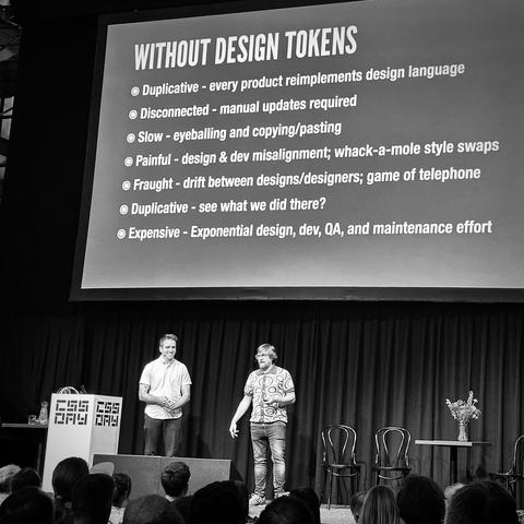 Ian and Brad on stage. Text-only slide:

WITHOUT DESIGN TOKENS
• Duplicative - every product reimplements design language
• Disconnected - manual updates required
• Slow - eyeballing and copying/pasting
• Painful - design & dev misalignment; whack-a-mole style swaps
• Fraught - drift between designs/designers; game of telephone
• Duplicative - see what we did there?
• Expensive - Exponential design, dev, QA, and maintenance effort