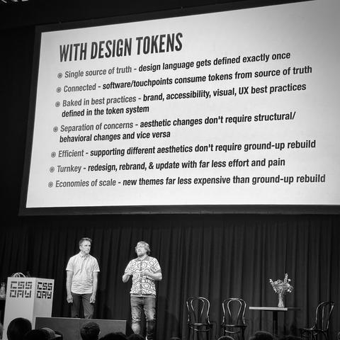 Ian and Brad on stage. Text-only slide:

WITH DESIGN TOKENS
• Single source of truth - design language gets defined exactly once
• Connected - software/touchpoints consume tokens from source of truth
• Baked in best practices - brand, accessibility, visual, UX best practices defined in the token system
• Separation of concerns - aesthetic changes don't require structural/ behavioral changes and vice versa
• Efficient - supporting different aesthetics don't require ground-up rebuild
© Turnkey - redesign, rebrand, & update with far less effort and pain
• Economies of scale - new themes far less expensive than ground-up rebuild