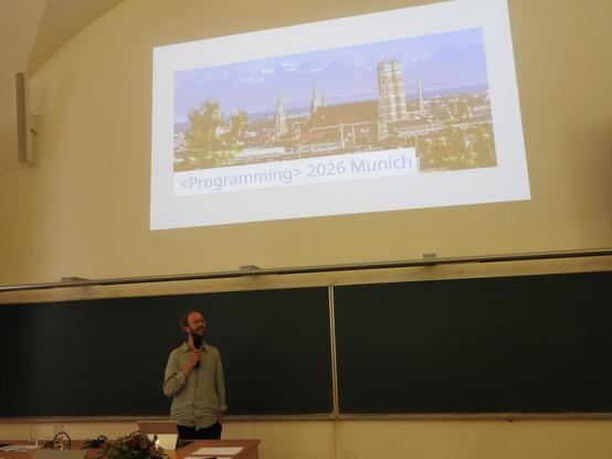 A photo of the announcement that <Programming> 2026 will take place in Munich. General chair of <Programming> 2025 Tomas Petricek is standing in the lower half of the picture holding a microphone. In the upper part is a projected slide with a photo of Munich. The slide says "<Programming> 2026 Munich"