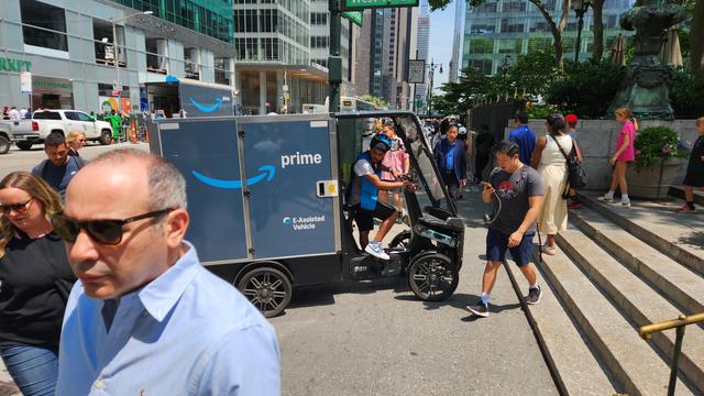 A four-wheeled Amazon "e-assisted vehicle " blocks a busy sidewalk outside of Bryant Park in Manhattan. An Amazon box truck is visible across the street in the background.