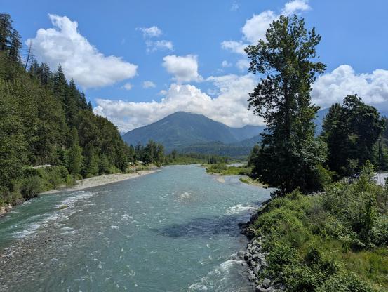 Im Vordergrund ist ein Fluss zu sehen, an beiden Ufern jeweils verschiedene Bäume, Büsche und Gras. Im Hintergrund ein grün bewachsener Berg und ein paar Wolken im sonst blauen Himmel.