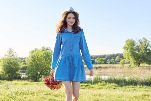 A beautiful young woman in a blue dress and white hat carrying a basket of strawberries near a pond.