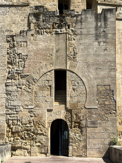 View of a historic wall featuring a blocked horseshoe-arched portal and a smaller, later doorway with a gate. The wall displays various construction phases with rubble stone, ashlar, and brickwork. 