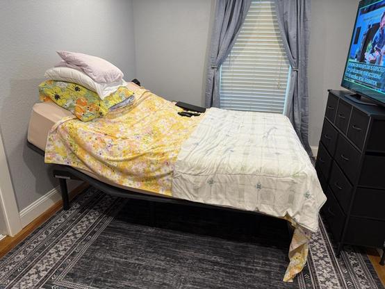 Small bedroom with a queen bed with the head tilited up, 4 pillows stacked, yellow floral sheets. A white comforter is folded across the foot of the bed. A black dresser is 6" away from the foot of the bed. It has a large TV on top of it that is level with the bed. The TV is on. A gray and black patterned rug extends from under the bed. The walls are light gray and the window has medium blue gray floor length curtains, pulled open in a swag. The blinds are closed.