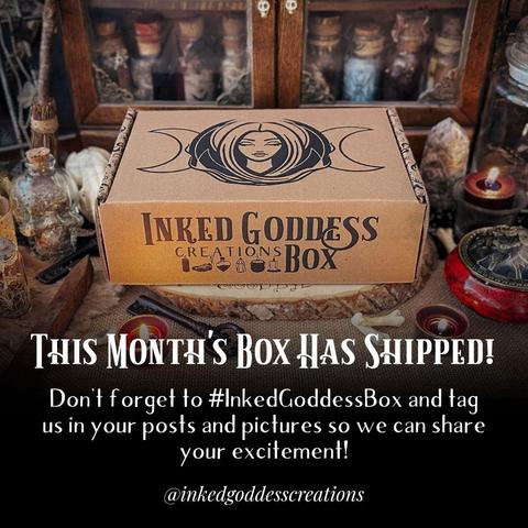 A rectangular brown cardboard box is set on a raw wood slice, surrounded by witchy items like gemstones, candles, and vials of herbs. The box features our exclusive brand packaging. The text across the top reads, "This Month's Box Has Shipped!" The Inked Goddess Creations Box is an exclusive product from Inked Goddess Creations.