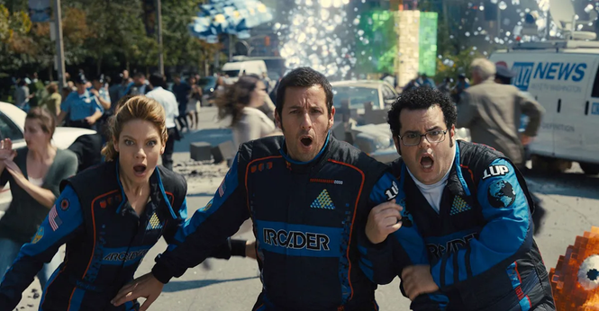 Cena do filme Pixels. Violet van Patten (Michelle Monaghan), Sam Brenner (Adam Sandler) e Ludlow Lamonsoff (Josh Gad) aparecem lado a lado, observando algo fora da imagem com expressões de espanto. Os três vestem macacões de corrida de carro. O cenário ao fundo mostra pessoas correndo e destruição, indicando perigo.