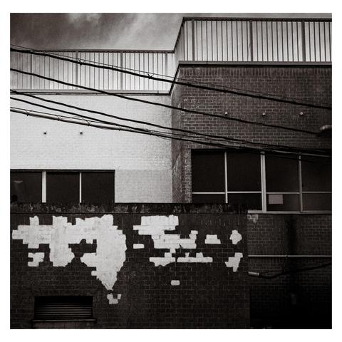 A tired brick wall in Nagoya puts on a performance of paint and indifference. Faded white rectangles trail off like a half-remembered promise, framed by taut overhead cables and mismatched architecture. Above, clean lines and rooftop fences suggest order.