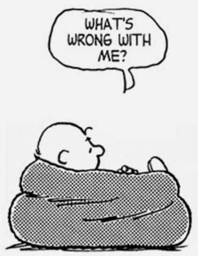 The image is a black and white cartoon featuring a character lying on a large, cushioned object. The character has a round head, a small nose, and a simple, content expression. The character is looking to the side, with one hand resting on the cushion. Above the character, there is a speech bubble containing the text "WHAT'S WRONG WITH ME?" The background is plain and white, emphasizing the character and the text. The style is reminiscent of classic comic strips, with simple lines and minimal detail.