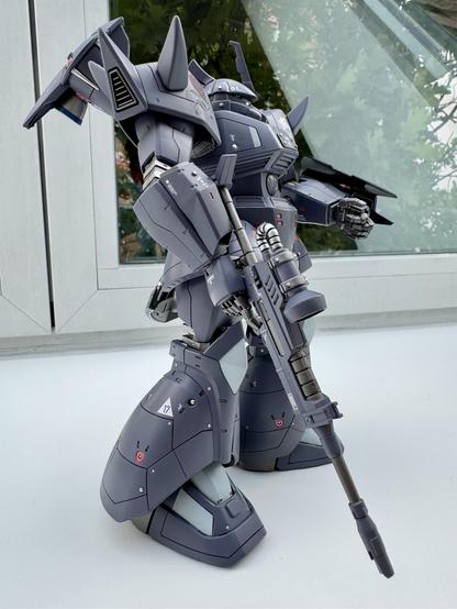 A detailed model of a futuristic robot figure stands in profile, featuring a predominantly gray color scheme with angular designs and various mechanical components. It is equipped with a large gun and has pronounced spikes on its shoulder armor. The background includes blurred greenery and a window. 