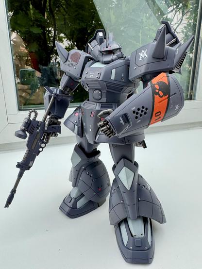 A detailed model of a robotic figure, featuring a dark gray and black color scheme, intricate mechanical parts, and a large gun in its right hand. The figure stands on a white surface with greenery reflected in the nearby glass.