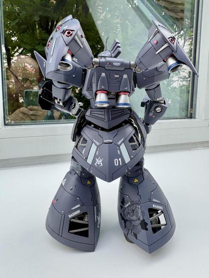 A detailed model of a mechanized robot is displayed from the rear, featuring gray armor, various mechanical details, and distinctive markings, including a design of a flower. The background includes reflections of greenery and a window.