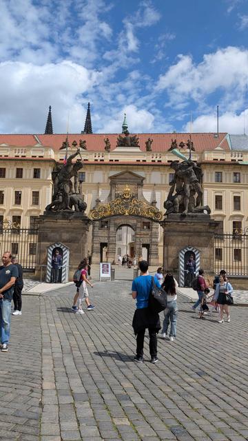 A photo of the gate near Prague Castle. Some people are standing in the foreground. In the middle of the photo is a gate with intricate decorations and two guards posted at the sides. There is an orange-ish building behind the gate. Behind the building, the tops of two antique castle towers can be seen.